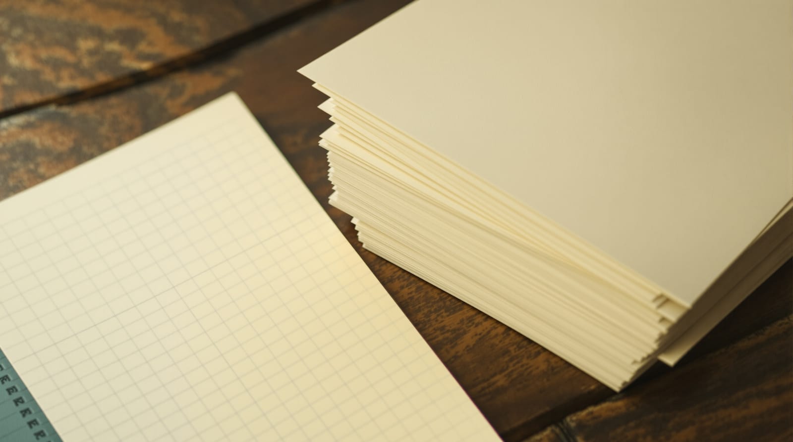 A stack of blank paper ballots beside a sheet of ruled grid paper on a wooden table